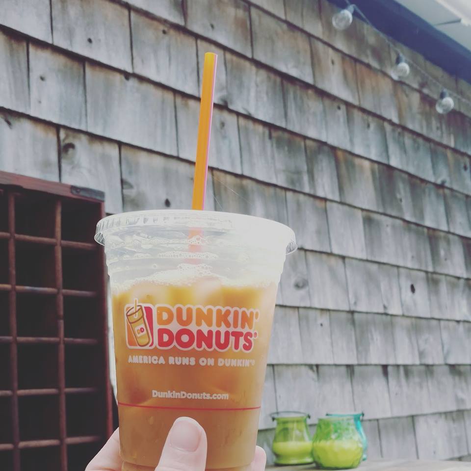 Cup of Dunkin' Donuts iced coffee being held up beside a house. The house has beach weathered shingles. May 23rd is Iced Coffee Day in Providence Rhode Island