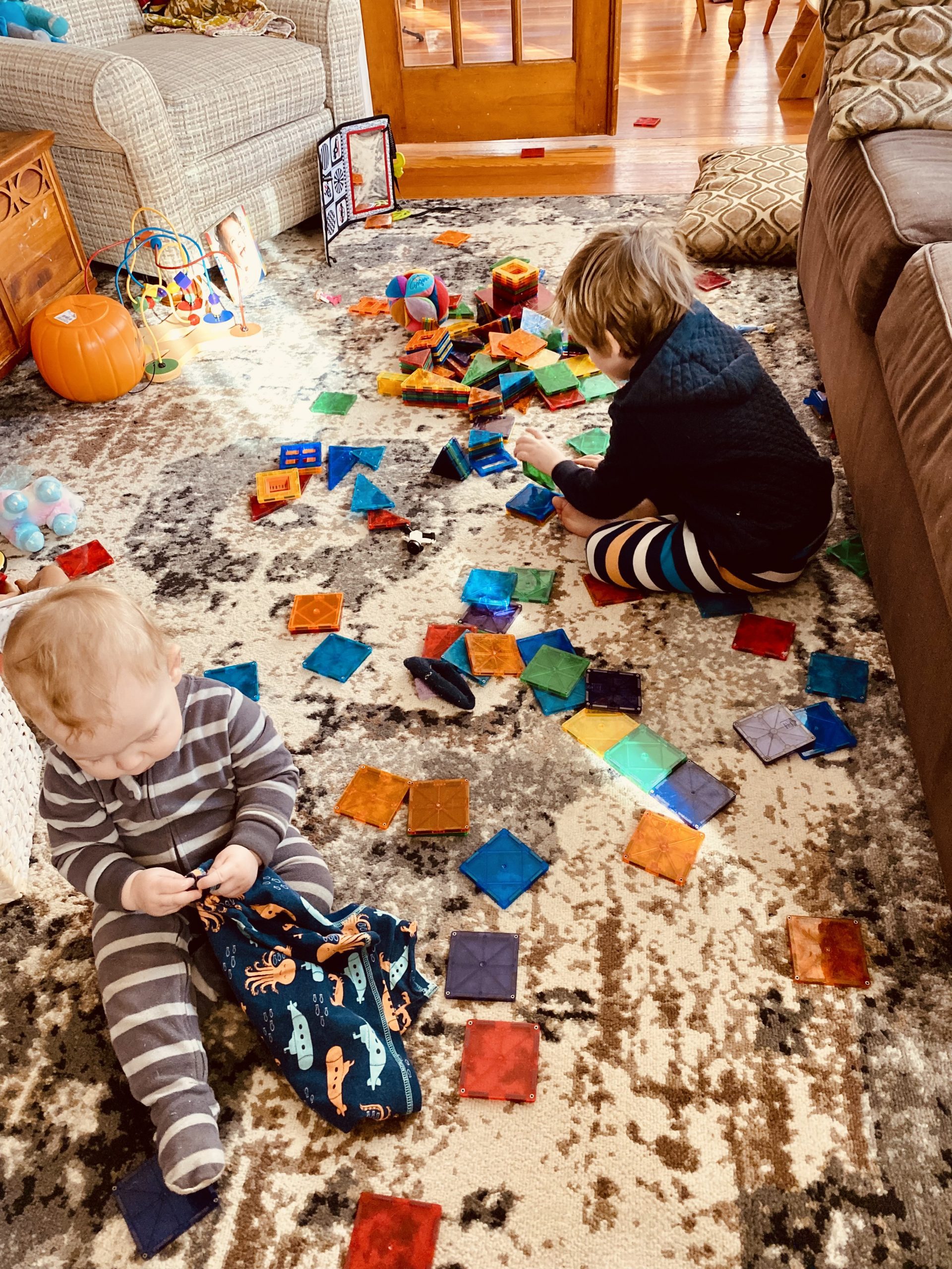 Two kids play on the floor surrounded by a mess of magnetic building tiles