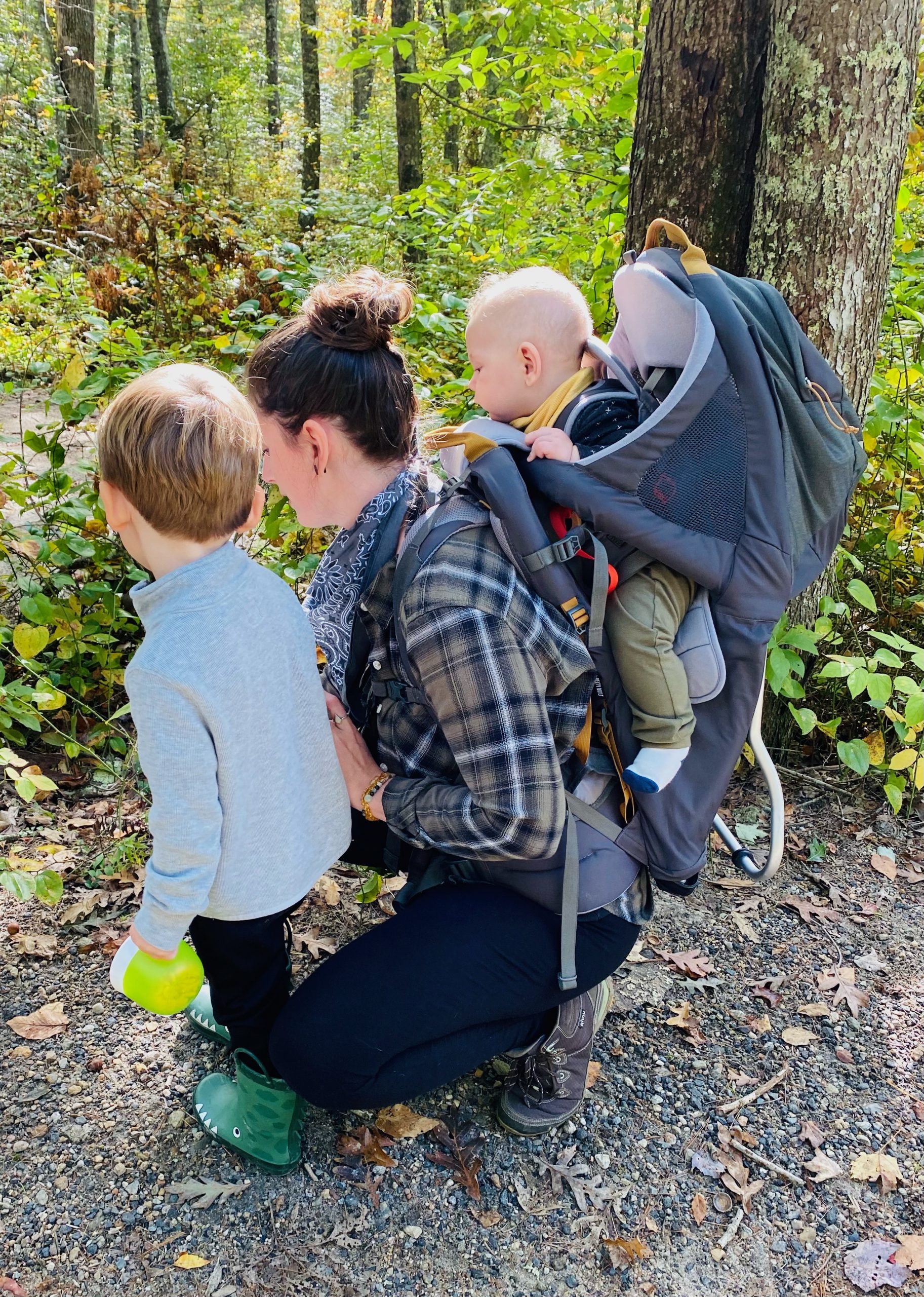 Mom on a hiking trail with a toddler by her side and a baby on her back in a backpack style carrier