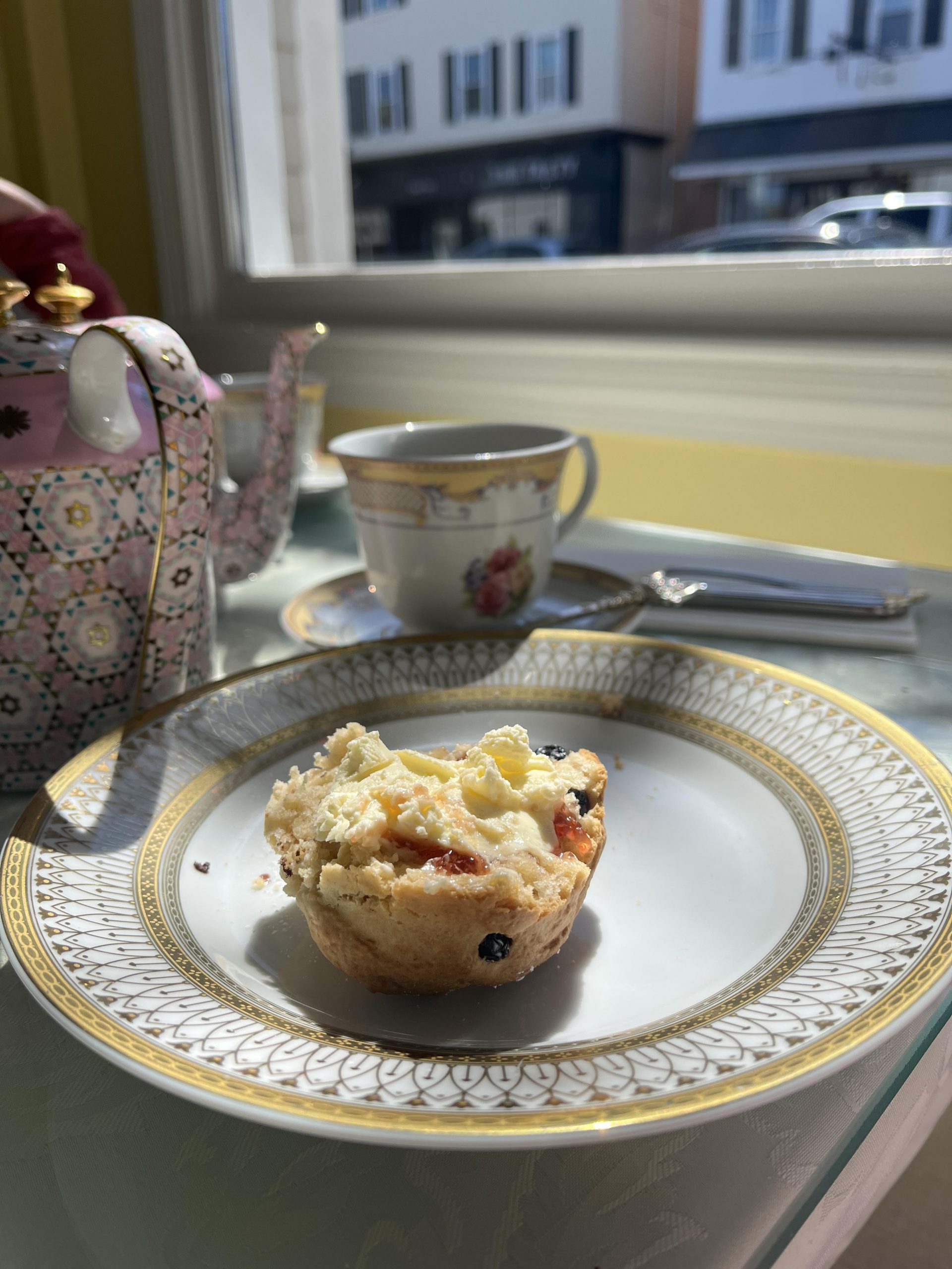 Half of a scone on a gold rimmed china plate is topped with jam and cream. A china tea cup with a gold rim and a pink rose design is behind the plate with a pink patterned teapot handle visible to the left of the cup.