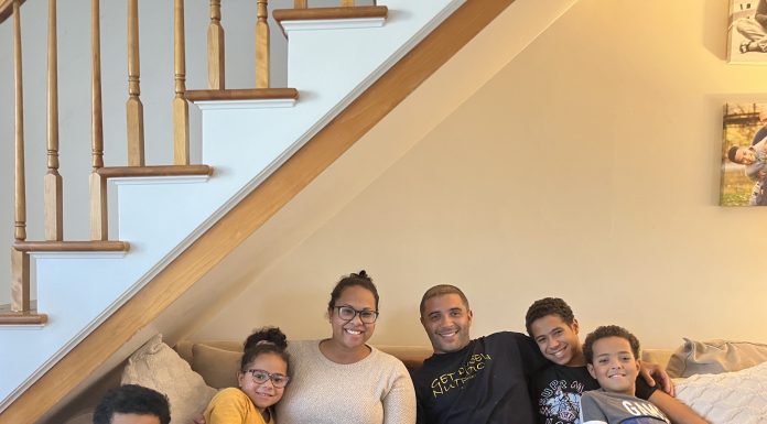 family of 6 sitting in living room together, all smiling, young black boy in red shirt, young black girl in yellow dress, black woman in cream sweater, black man in black shirt black teeanger in black shirt, young black boy in grey shirt