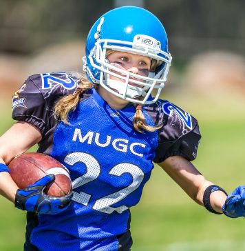 A girl wearing pigtails is dressed in full football uniform including helmet and mouthguard carries a football under her right arm.