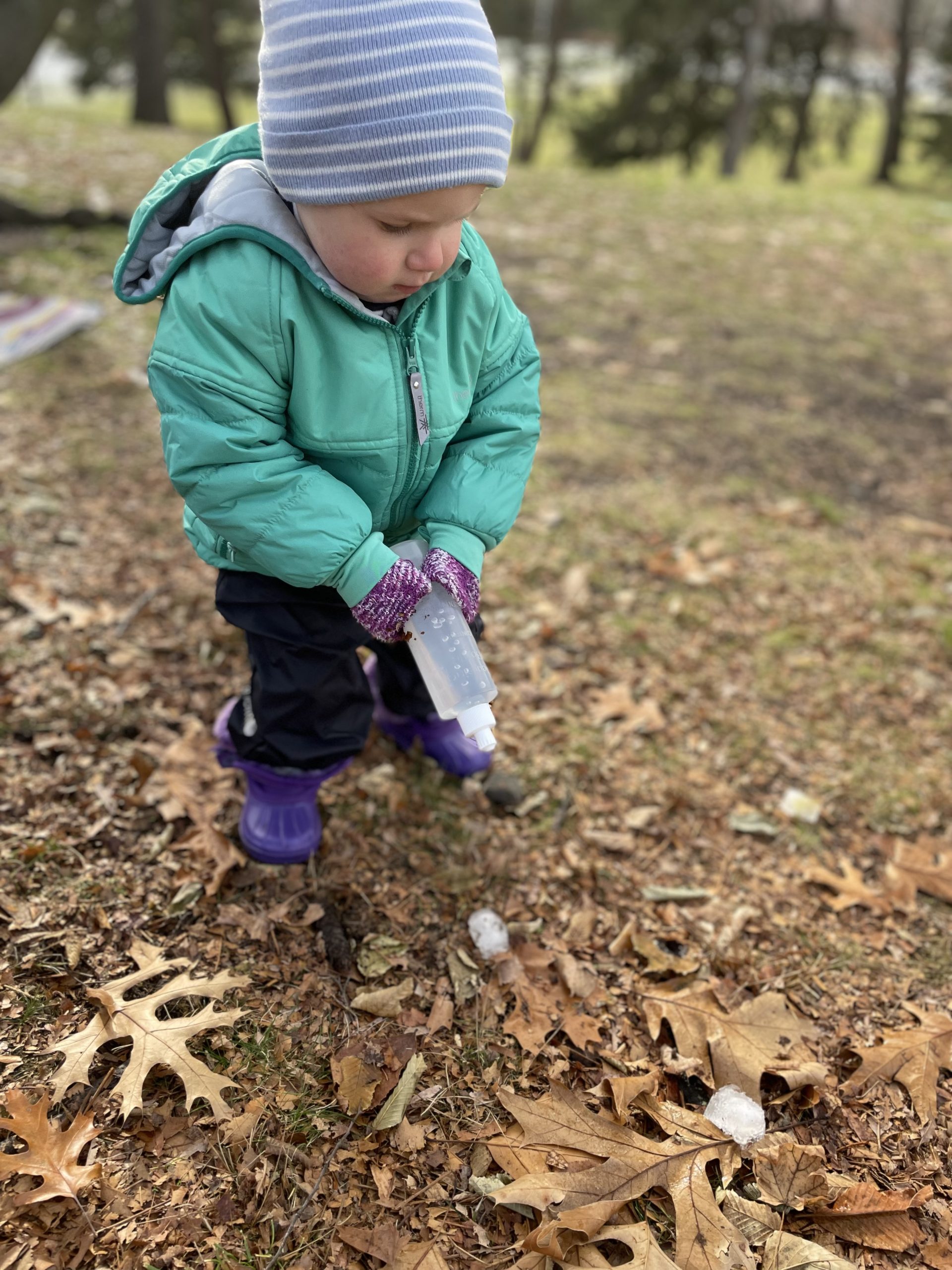 Toddler in hat and coat with a squeezy bottle of water. Cubes of ice are on the ground among the leaves.