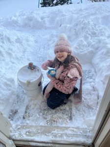girl in snow gear sits in the snow with a bucket full of snow and dolls in the bucket, She holds a bowl of snow and a spoon